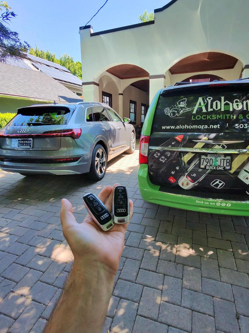 A person holds two Audi car keys in front of a silver Audi SUV parked in a driveway, with a green locksmith van labeled "Alohomora Locksmith & Garage Door" nearby.