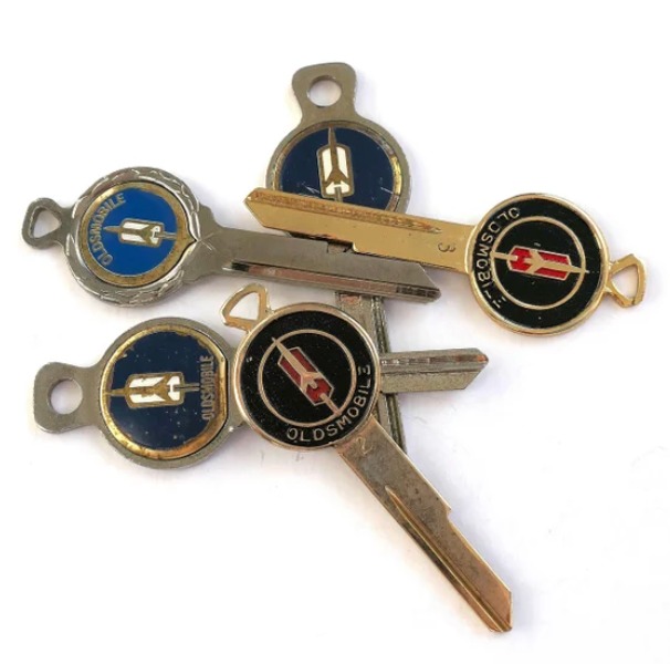 Four vintage metal car keys with circular Oldsmobile logo emblems on the heads, featuring blue, black, and gold colors, arranged in a fan shape on a white background.