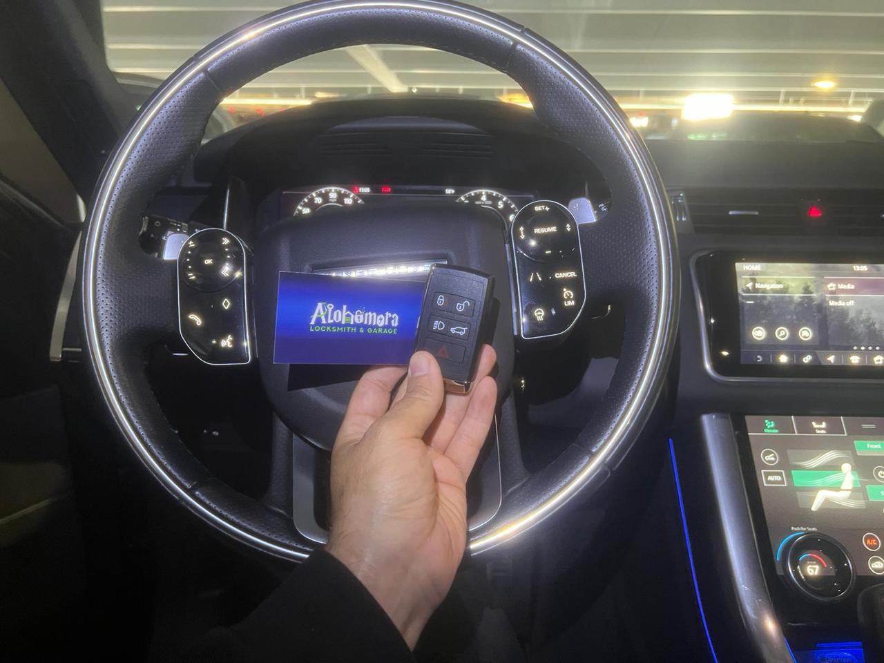 A person holds a car key fob and a blue card labeled "Alohimiora" in front of a steering wheel inside a modern car, with dashboard lights and infotainment screens visible.
