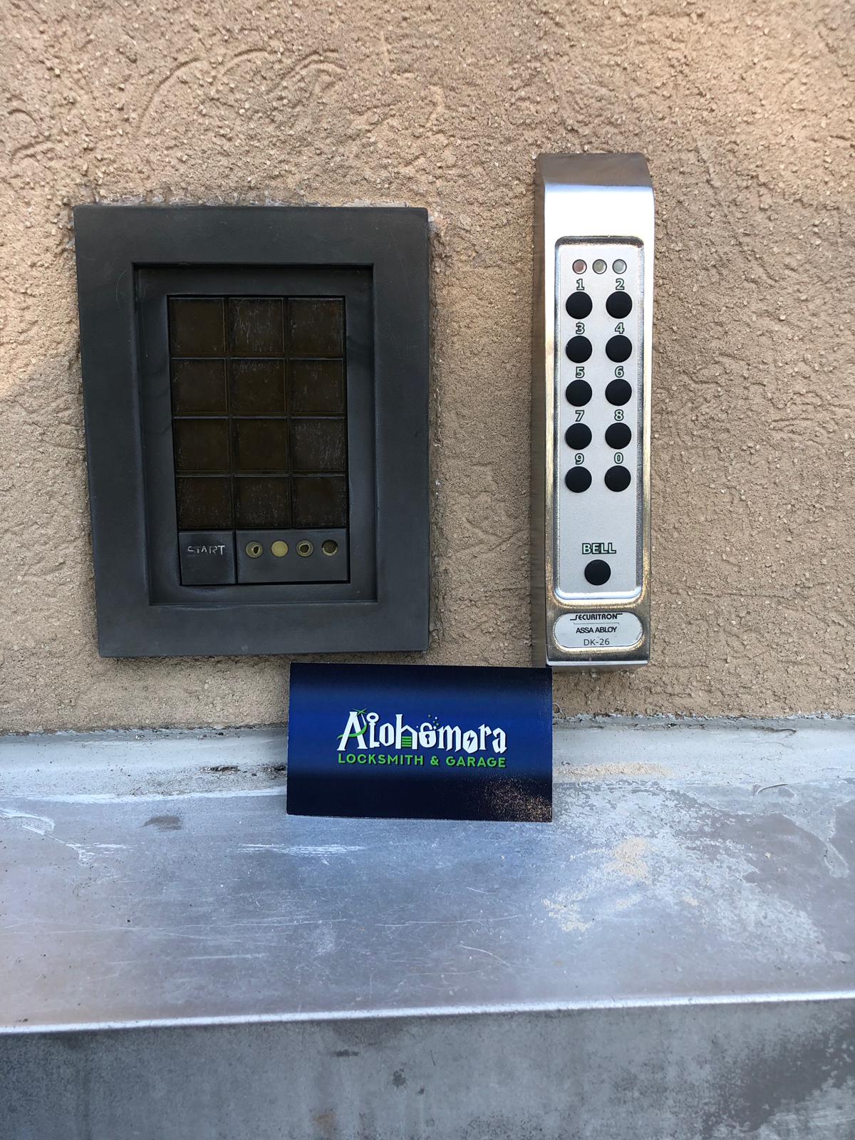 WhatsApp Image 2019-08-15 at 19.23.32 A keypad lock and a black square security panel are mounted on a beige wall. Below them is a blue business card for “Alohomora Locksmith & Garage.”.