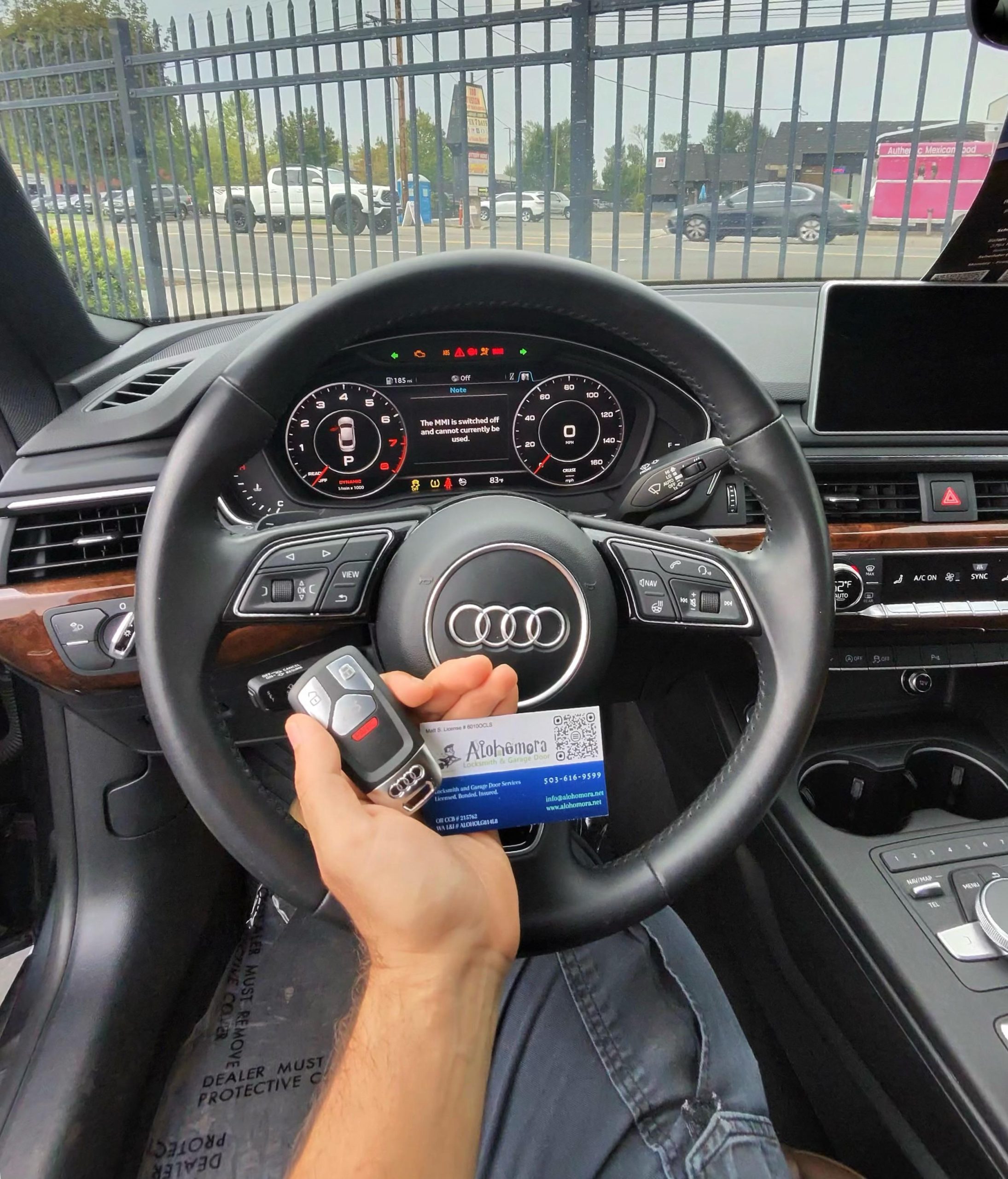 A person sitting in an Audi car holds a key fob and a business card in front of the steering wheel. The dashboard displays digital gauges, and parked cars can be seen through the windshield.
